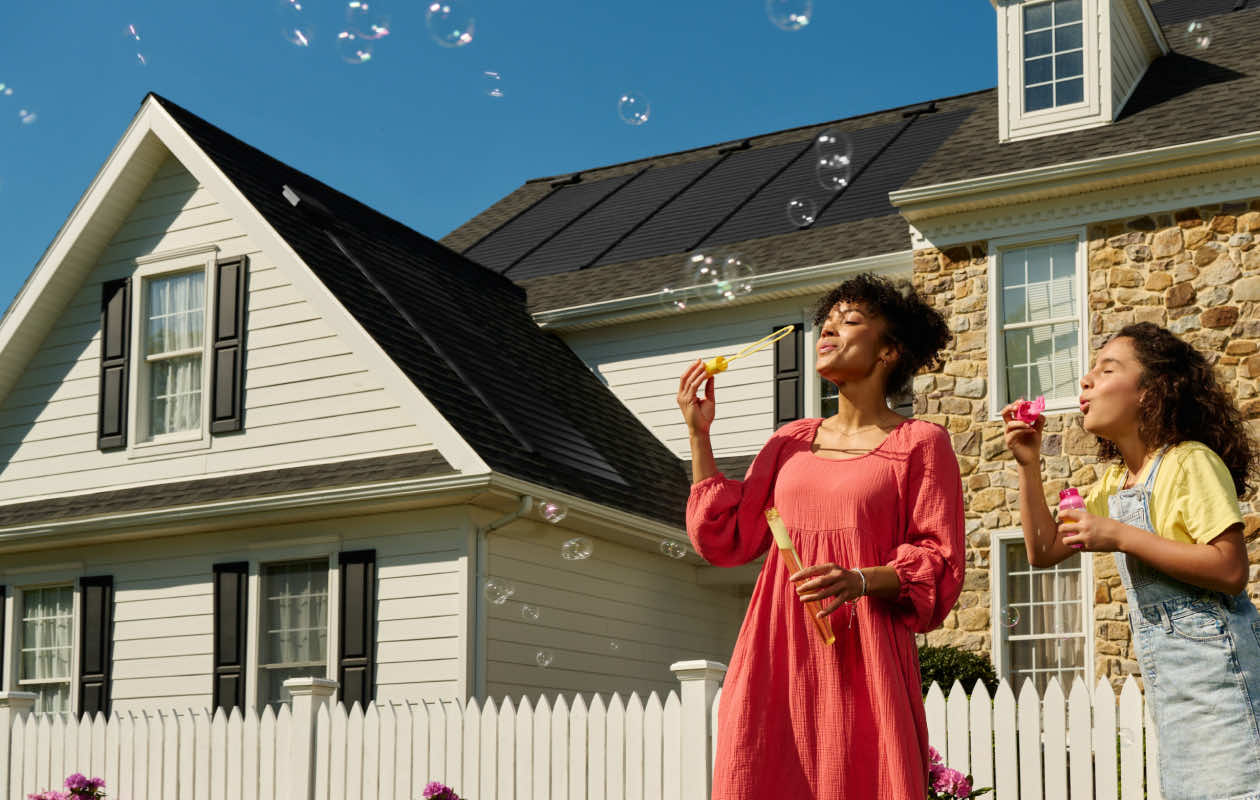 A mother and daughter blowing bubbles in front of a house with a solar roof and a white picket fence.