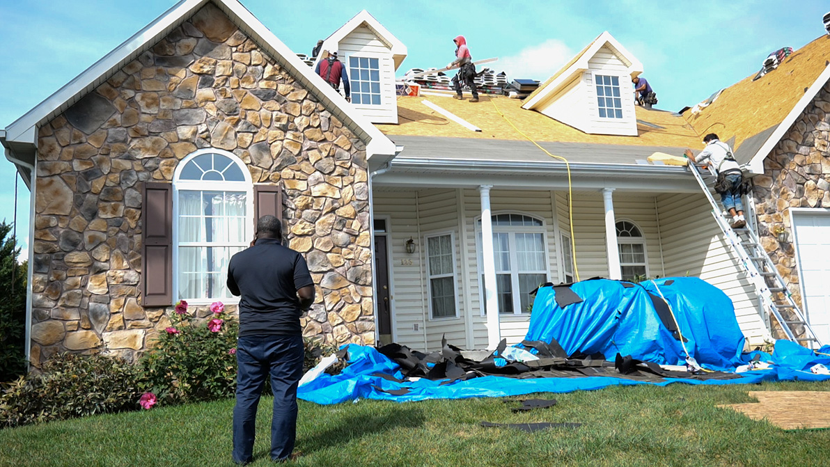 A man stands in front of a suburban house supervising a roofing crew working on the roof replacement. Several workers are on the roof with tools and materials, while tarps and debris cover the yard.