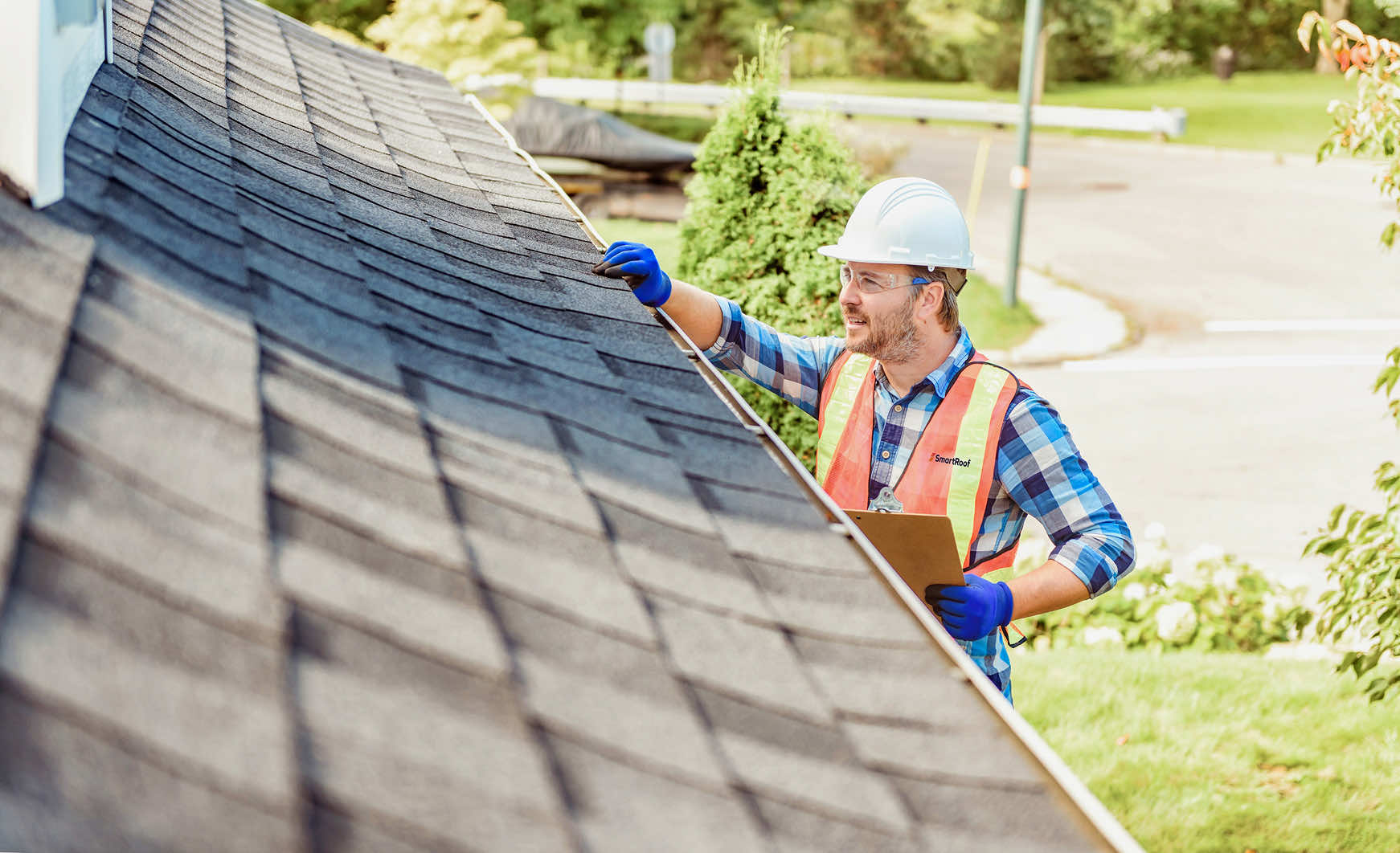A roofing worker inspecting shingles on a sloped roof with a clipboard.